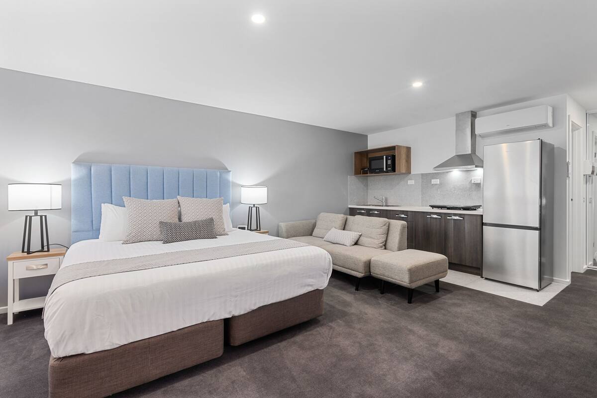 A modern hotel room with a large bed, a sofa, a kitchenette, and a separate bathroom.