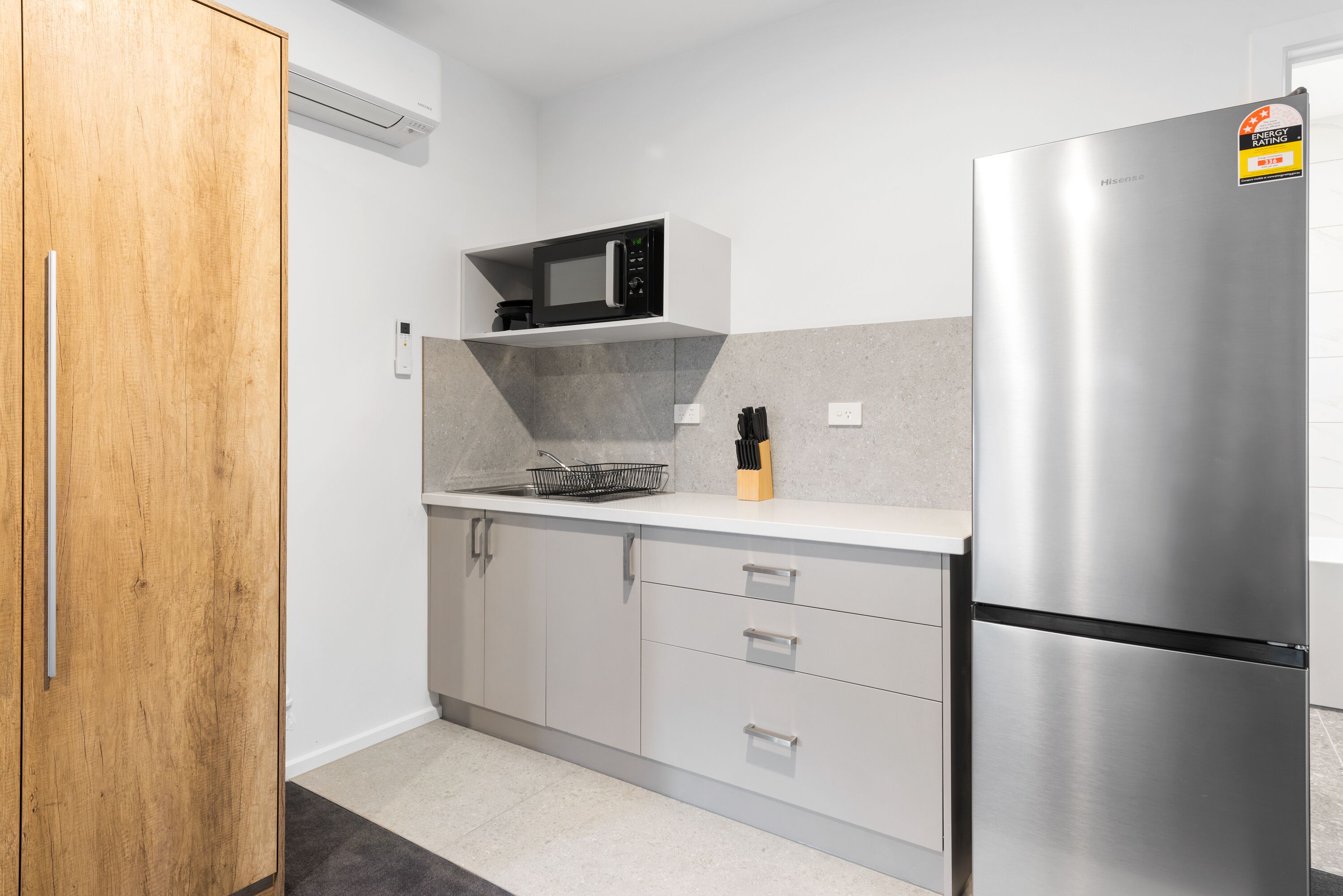 A modern kitchen with a stainless steel refrigerator, a microwave on a white shelf, and a wooden door.