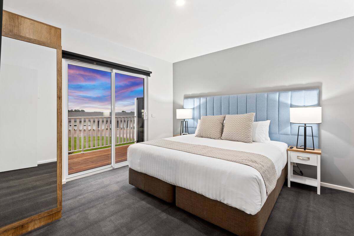 A modern bedroom with a large bed, a nightstand, a sliding glass door leading to a balcony, and a view of the sky.