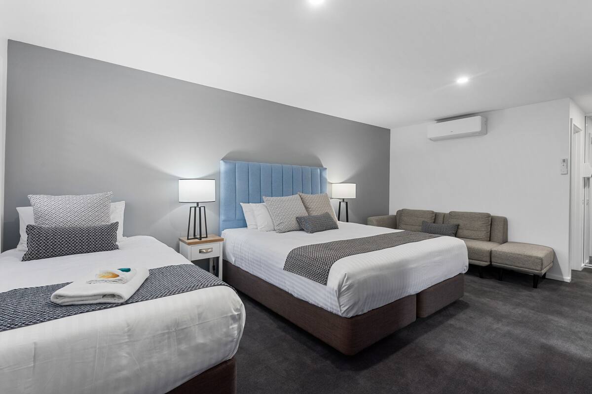 A modern hotel room with two beds, a sofa, and a nightstand with lamps.