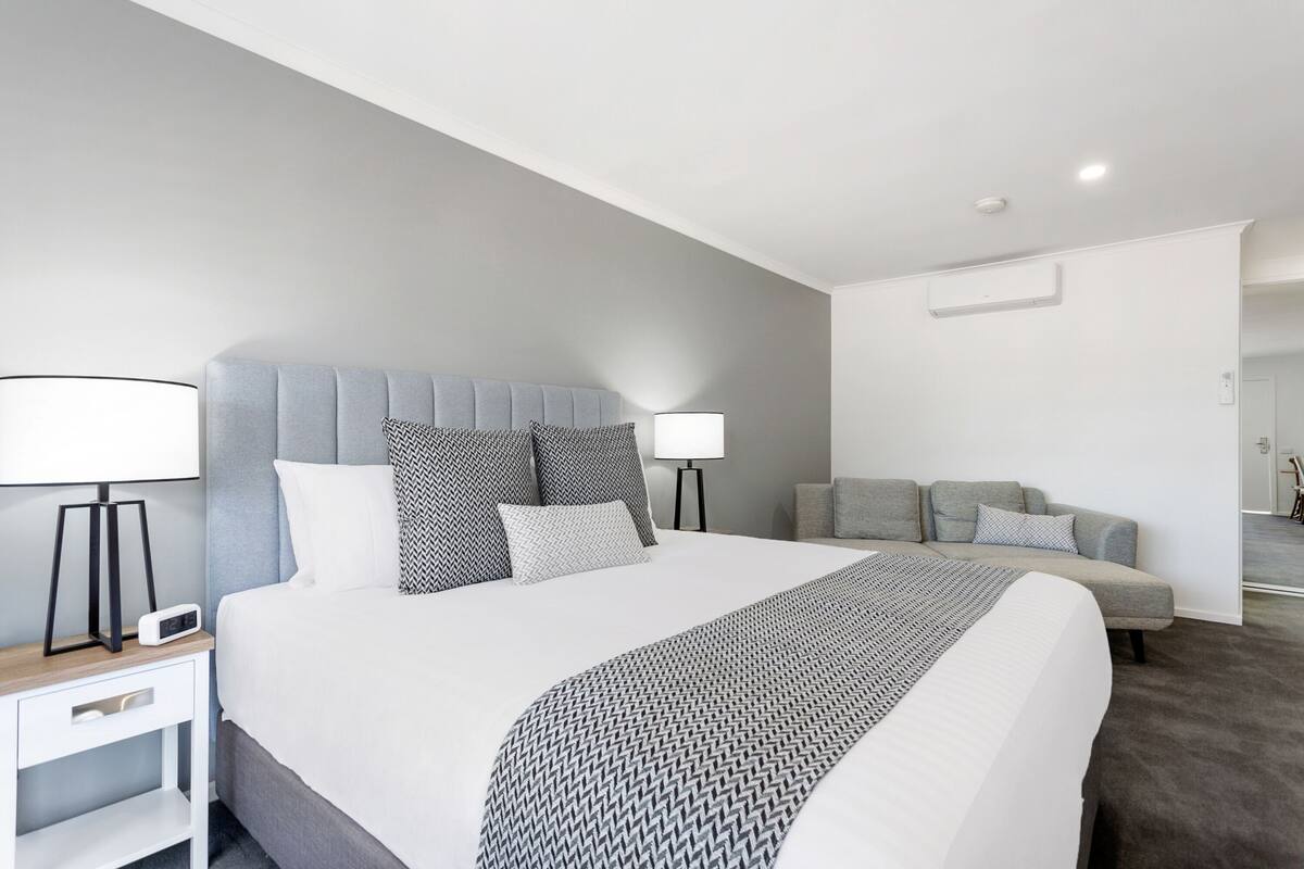 A modern hotel room with a large bed, two bedside tables, a grey sofa, and a small table with a lamp.