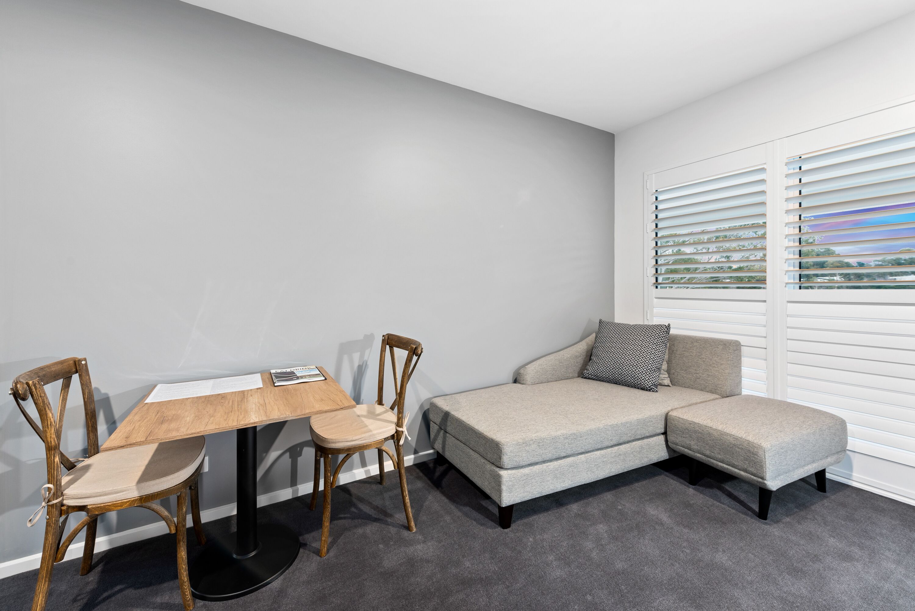 Une pièce moderne avec un canapé gris, une table à manger en bois et deux chaises, ainsi qu’une fenêtre avec des stores.