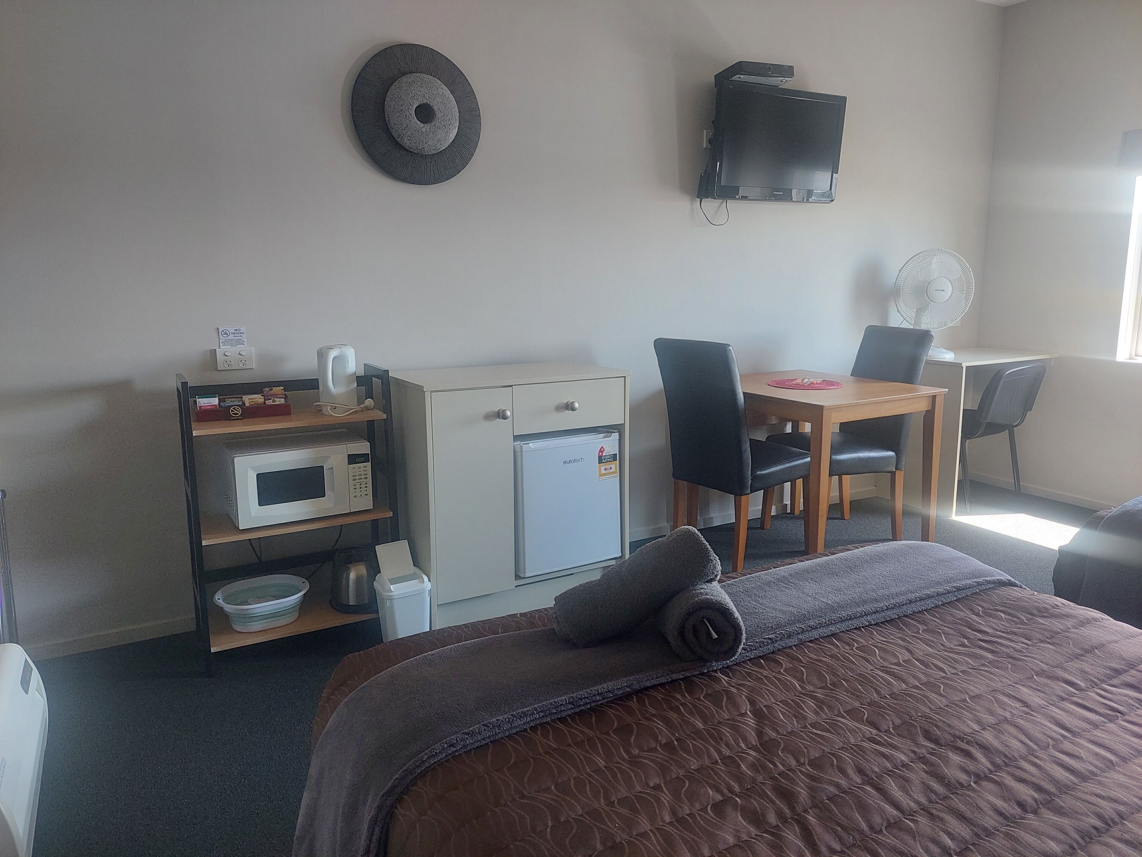 A hotel room with a bed, a desk, a microwave, a refrigerator, a chair, and a wall-mounted TV.