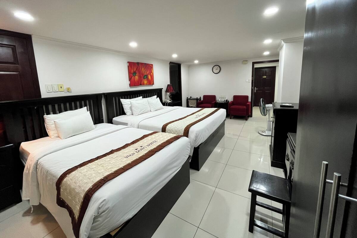 A hotel room with two beds, a desk, a chair, a clock, and a red flower painting on the wall.