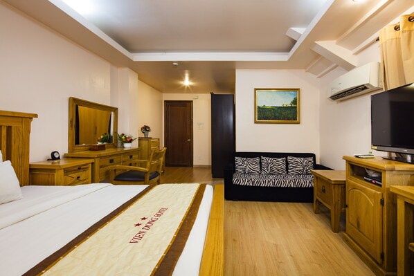 A hotel room with a bed, a desk, a chair, a sofa, a television, and an air conditioning unit.