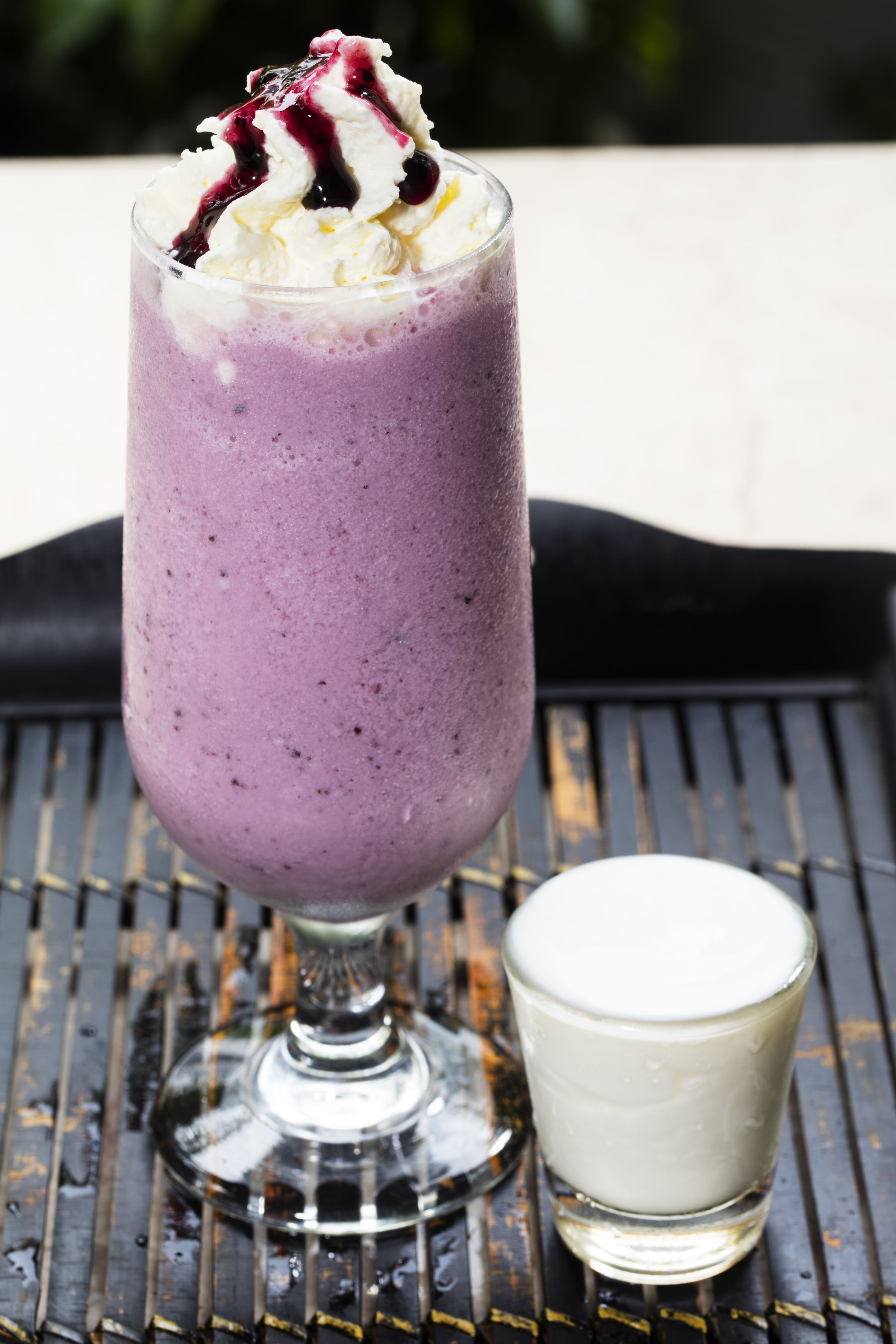A glass of purple smoothie with whipped cream and a berry drizzle, placed on a metal surface.