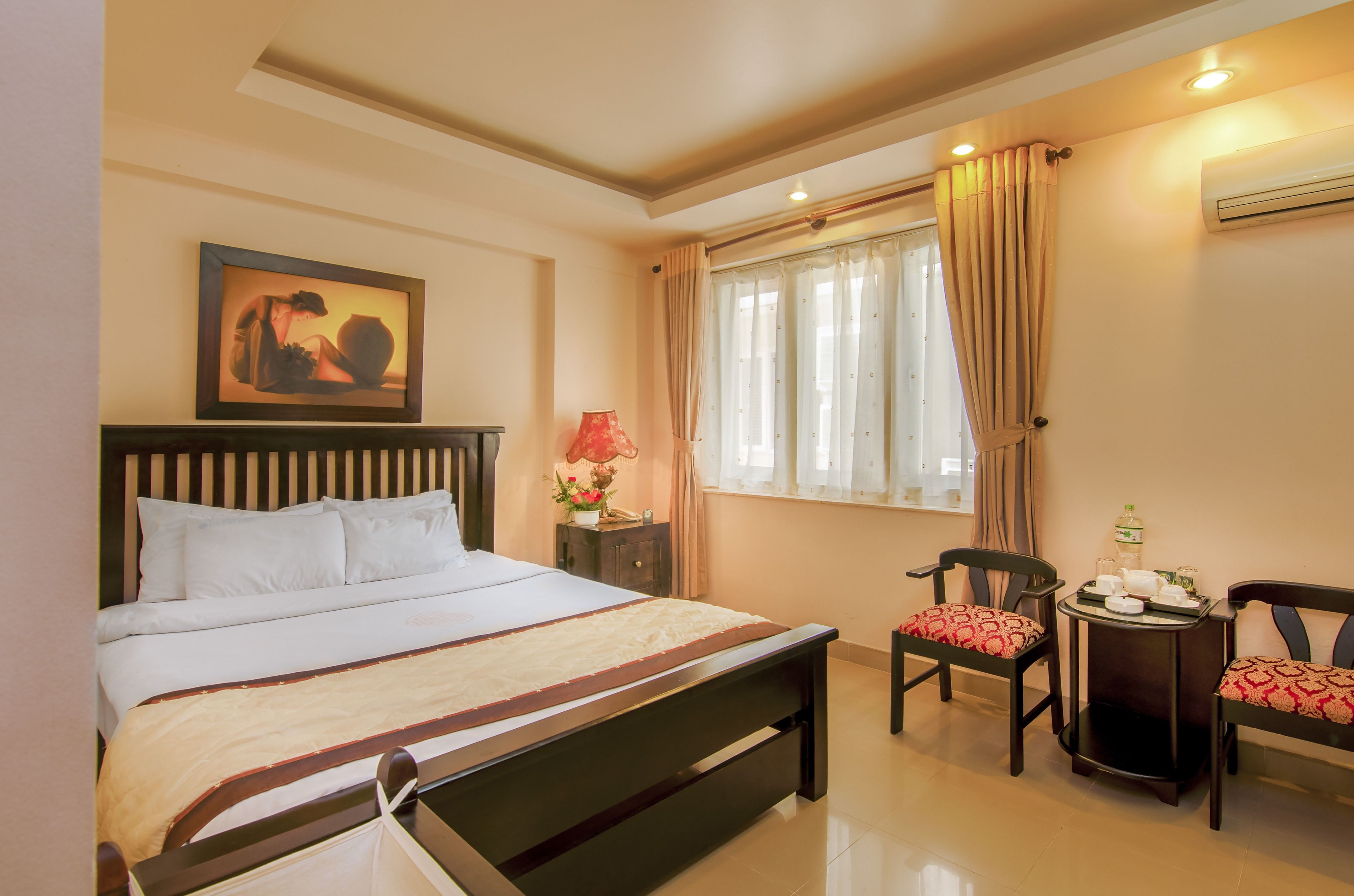 Une chambre d’hôtel bien rangée, avec un lit, une table de chevet, une lampe, des chaises et une fenêtre avec des rideaux.