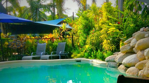 A pool area with lounge chairs, a table with a glass and a plant, and a stack of rocks. - Mission Reef Resort (Wongaling Beach)