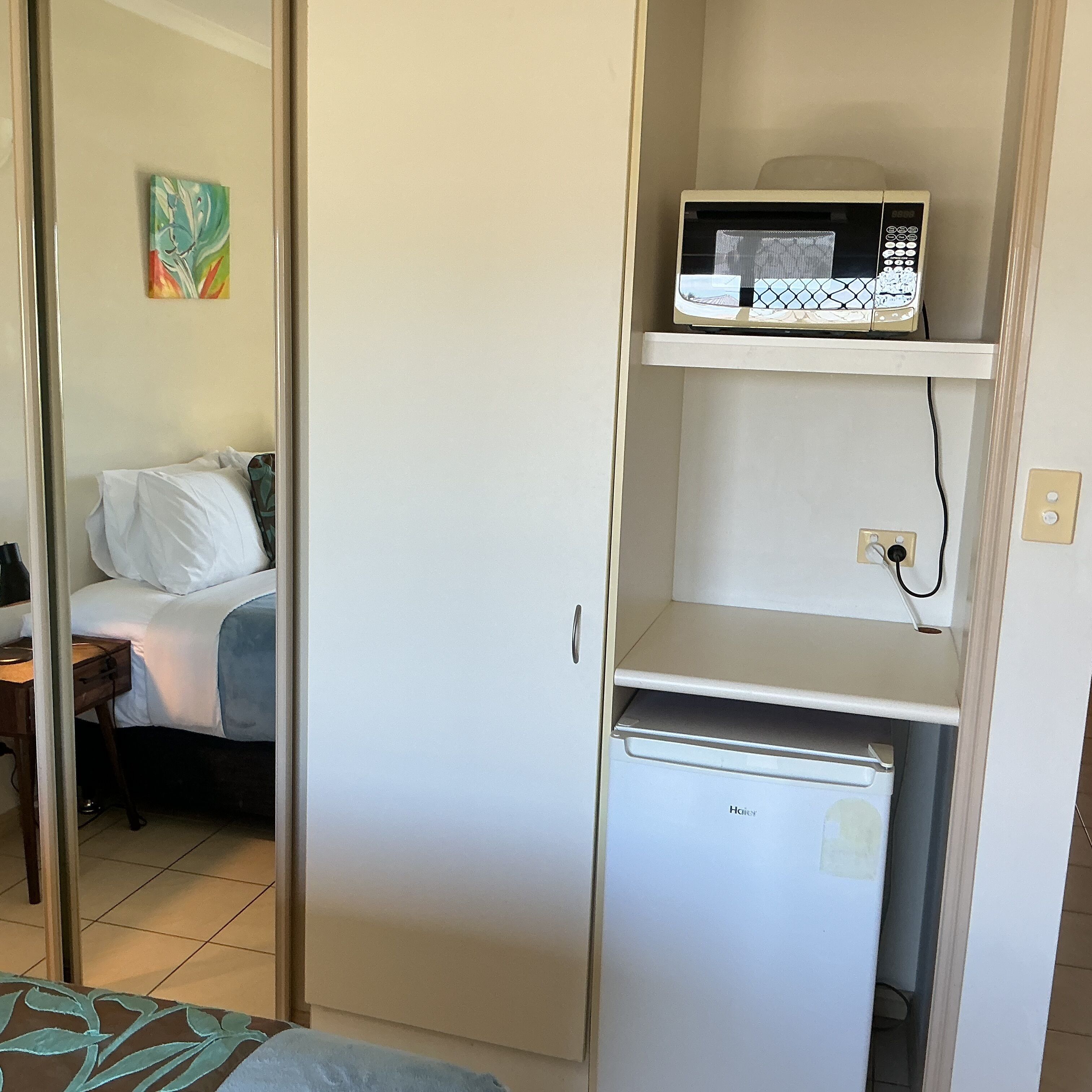 Habitación compacta con cama, microondas en un estante y un refrigerador pequeño.