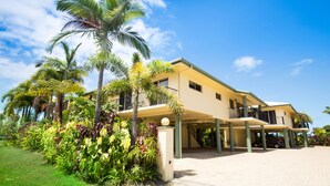 A two-story building with a covered parking area and a well-maintained garden with various plants and a palm tree. - Mission Reef Resort (Wongaling Beach)