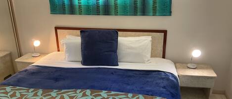 Una cama bien hecha con una colcha azul y blanca, dos almohadas blancas y una almohada decorativa azul oscuro. Hay dos lámparas de noche a cada lado de la cama.