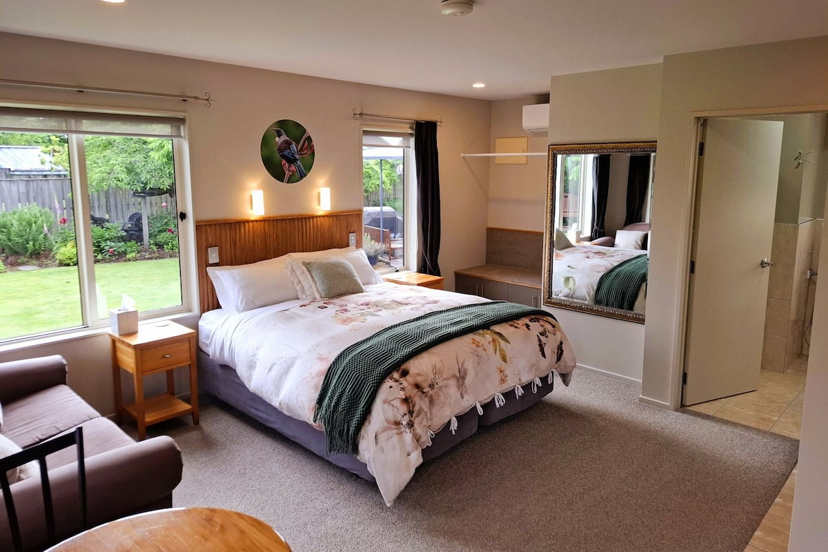 Une chambre avec un grand lit, une table de chevet, une chaise, une petite table, un miroir et une vue sur le jardin.