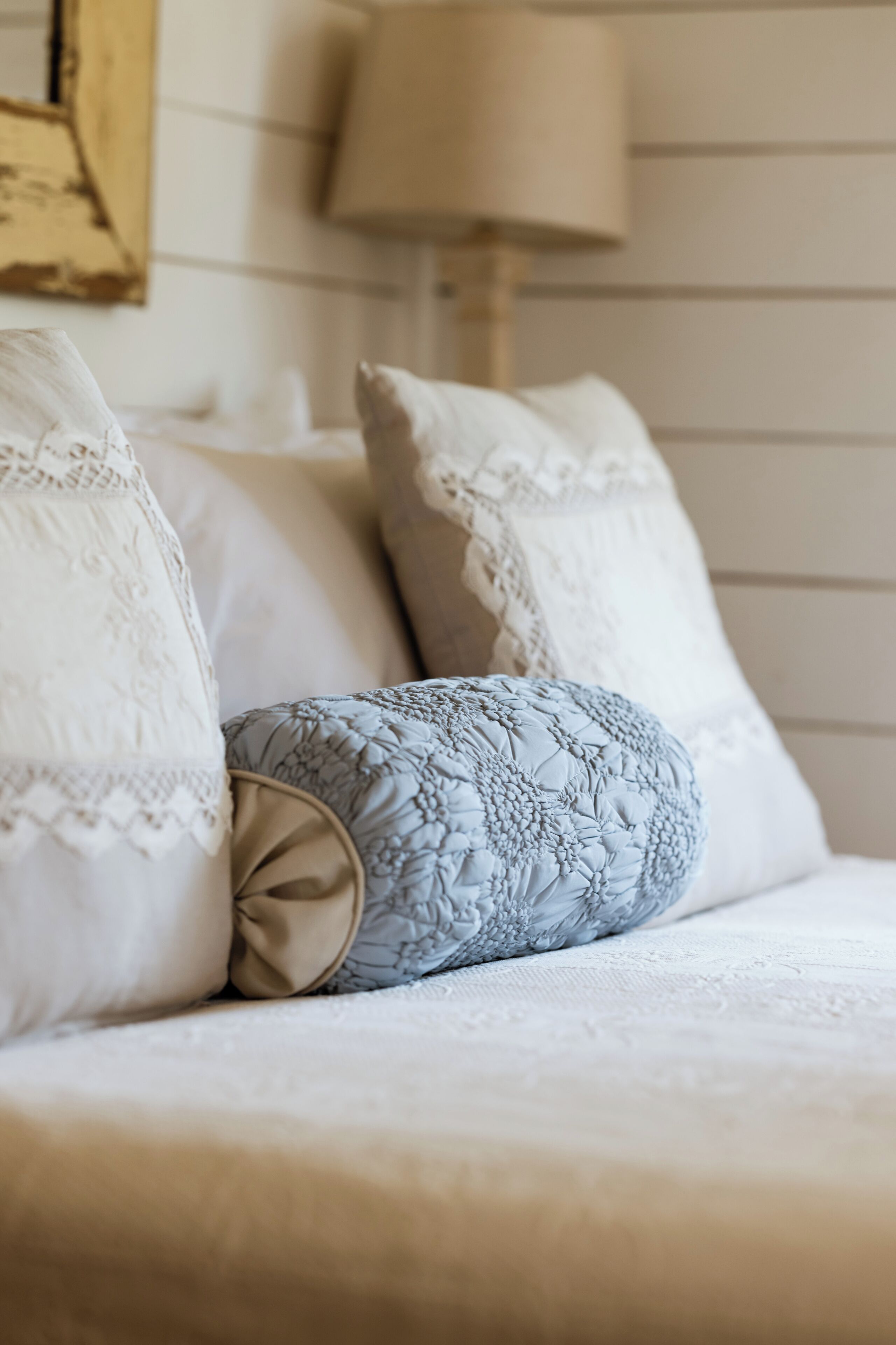 A bed with white and patterned pillows, a beige throw, and a lamp in the background.