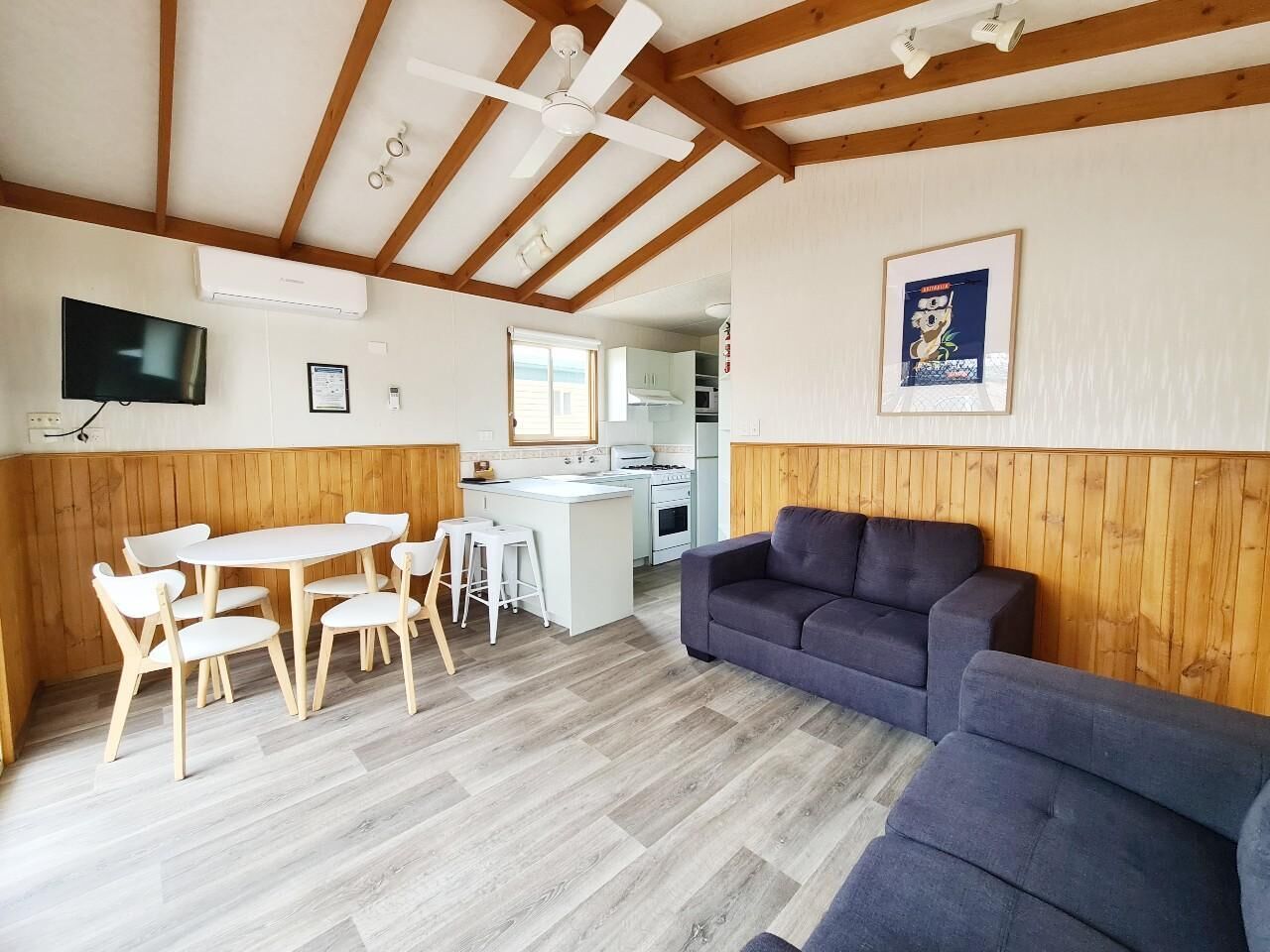 A modern kitchen with white cabinets and a central island, a dining area with a white table and chairs, and a living area with a dark-colored sofa.