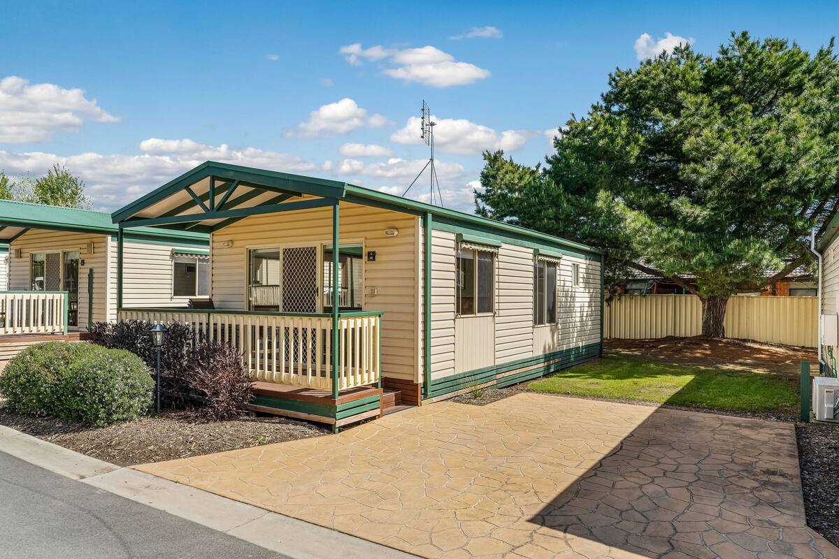 A mobile home with a covered porch, a fenced yard, and a large tree.
