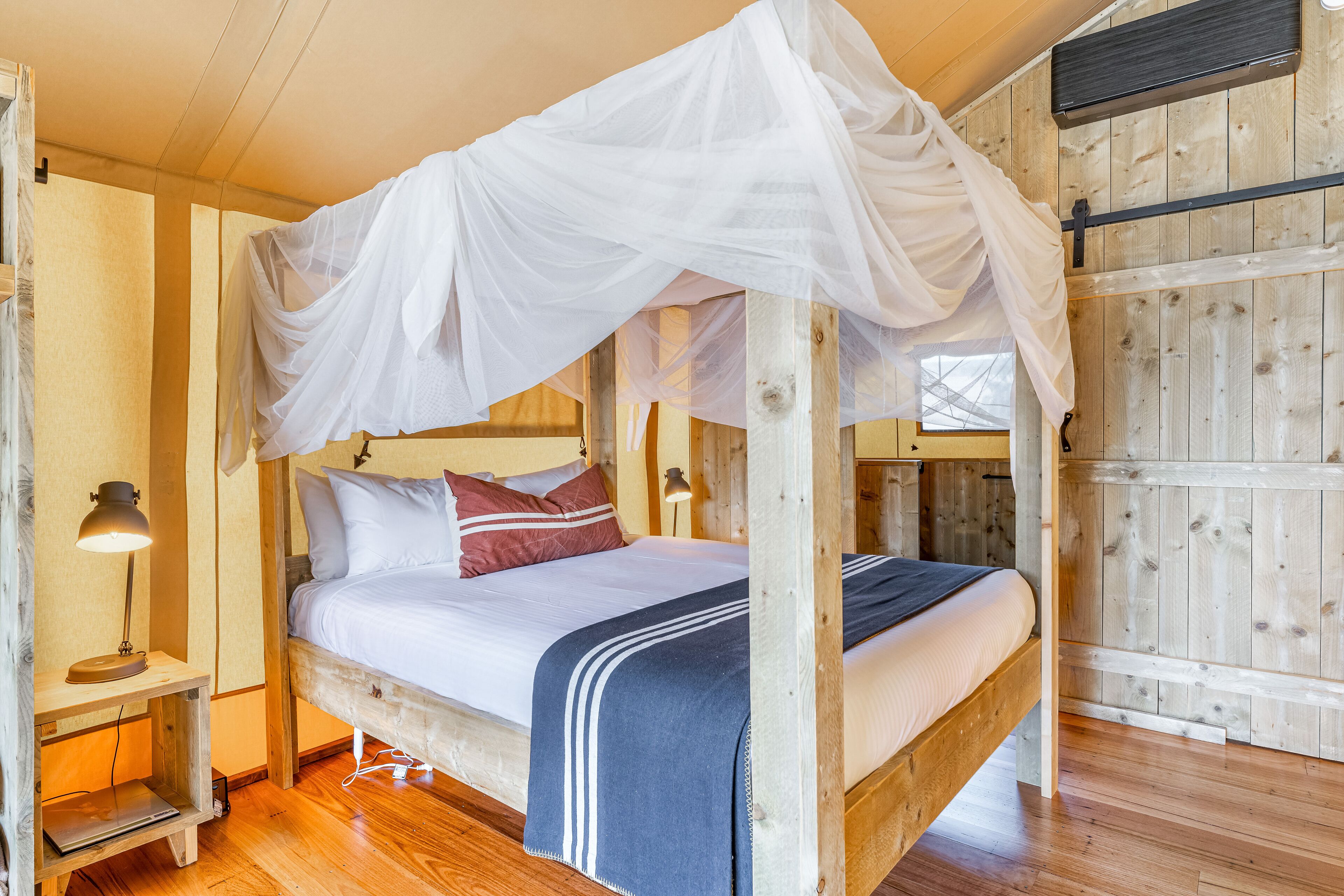 A wooden four-poster bed with a white canopy, a bedside table with a lamp, and a laptop on a shelf.