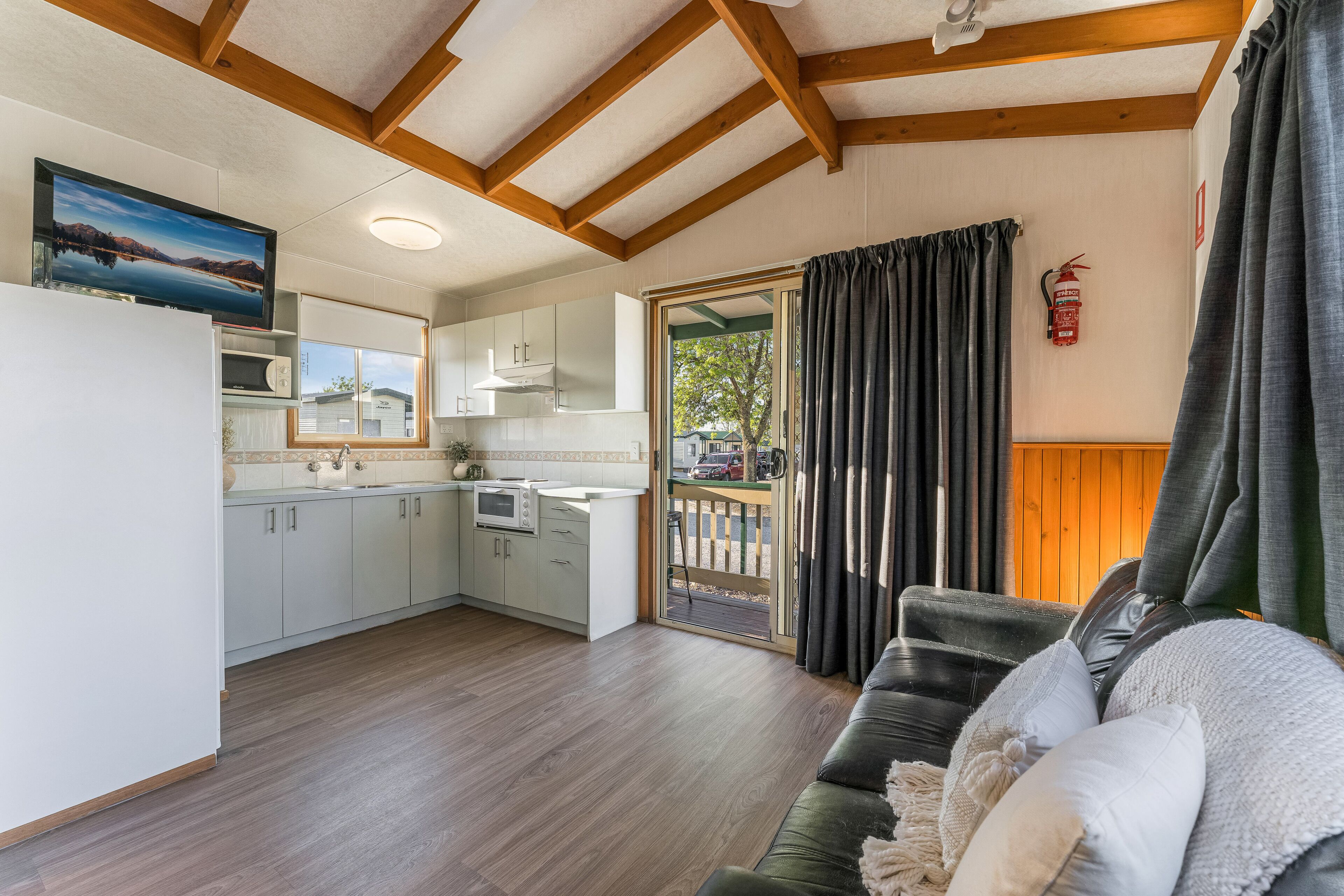 Une cuisine moderne avec des meubles blancs et une grande fenêtre, un salon avec un canapé vert foncé et une cheminée, et un balcon offrant une vue sur un immeuble et des arbres.