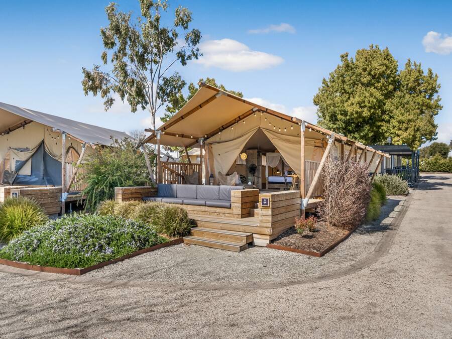 A caravan-style accommodation with a covered outdoor seating area, a gravel driveway, and surrounding vegetation.