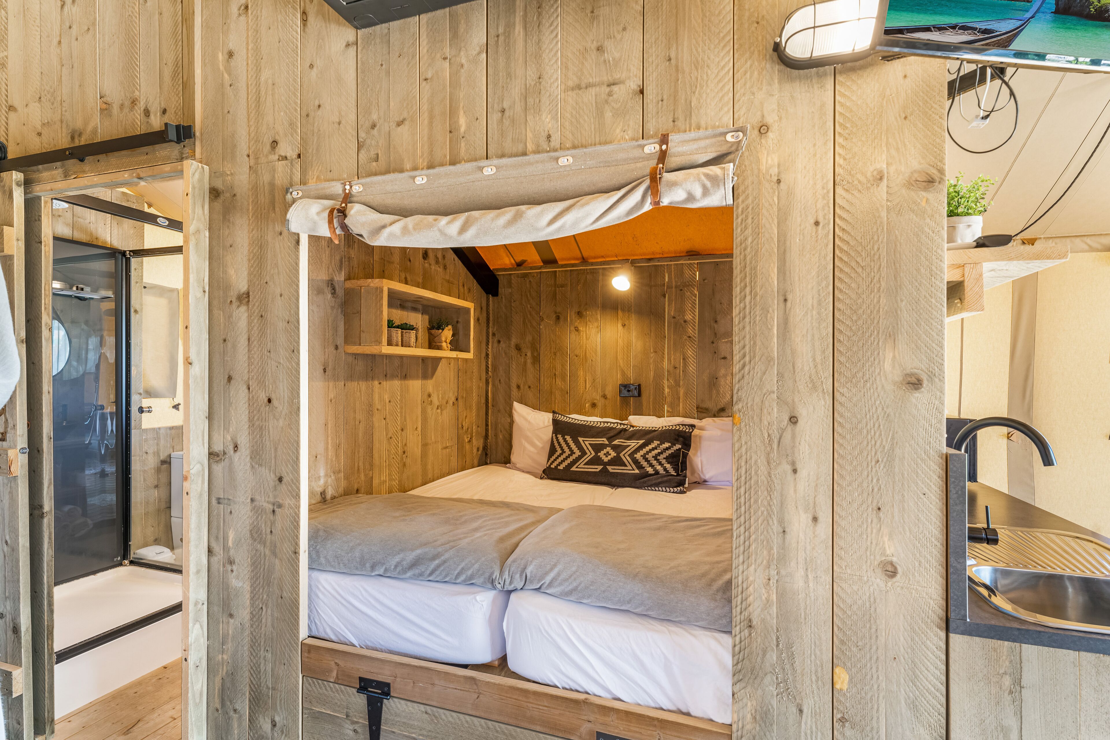A wooden cabin interior with a bed, a small shelf with plants, and a bathroom with a shower.