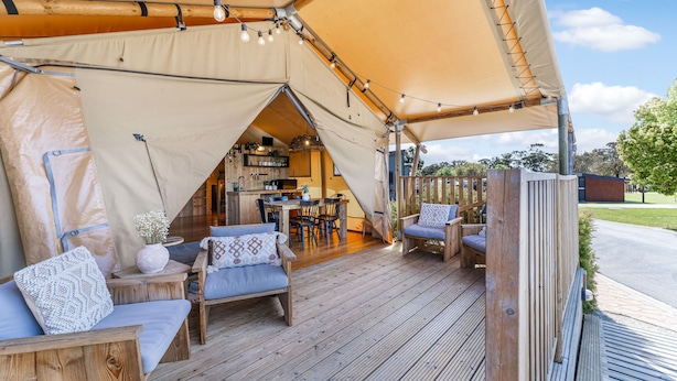Un área al aire libre con carpas, mobiliario de madera, una mesa de comedor y una cocina visible en el interior.