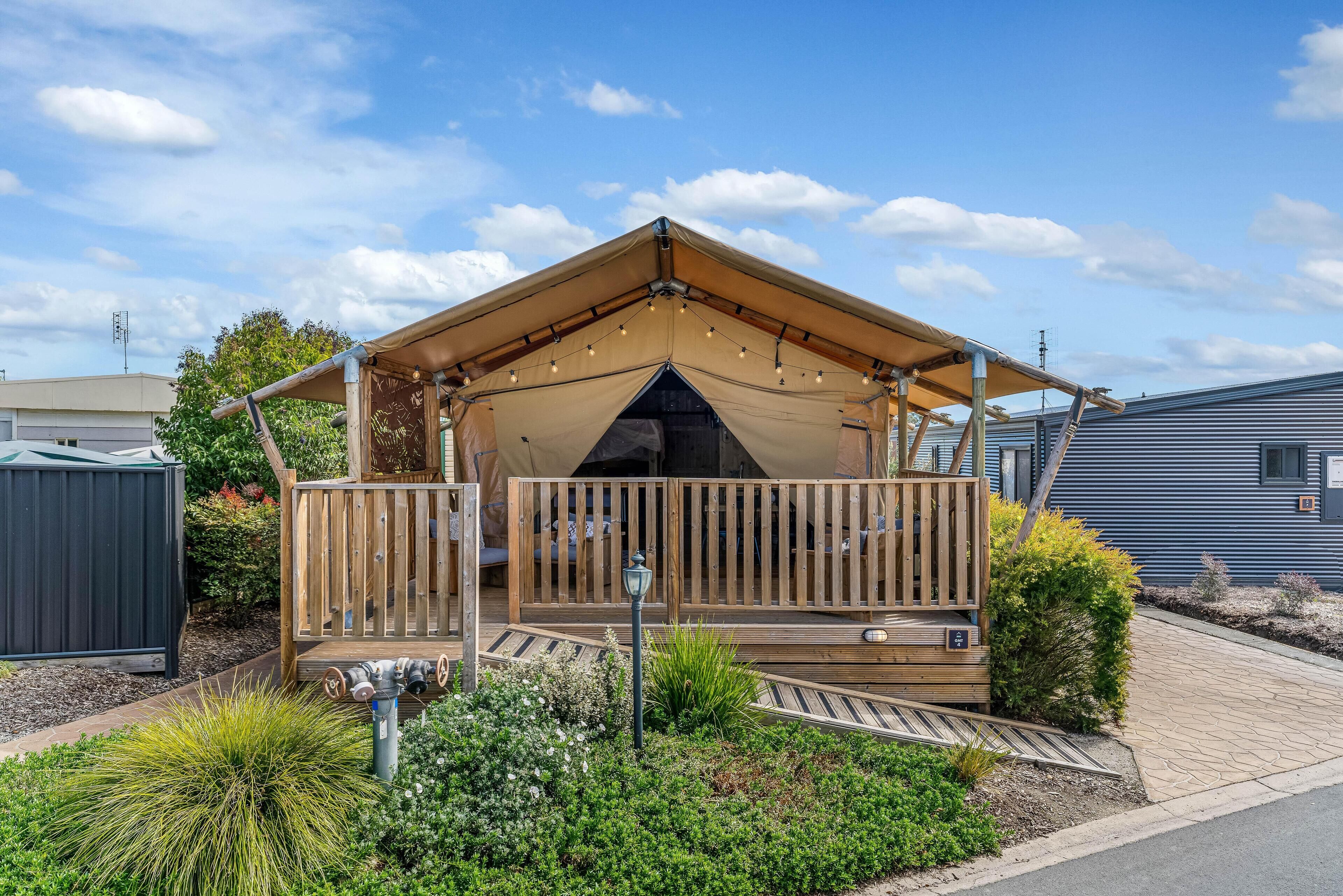 Une terrasse en bois dotée d’un espace salon couvert, entourée de parterres aménagés et agrémentée d’un petit luminaire extérieur.