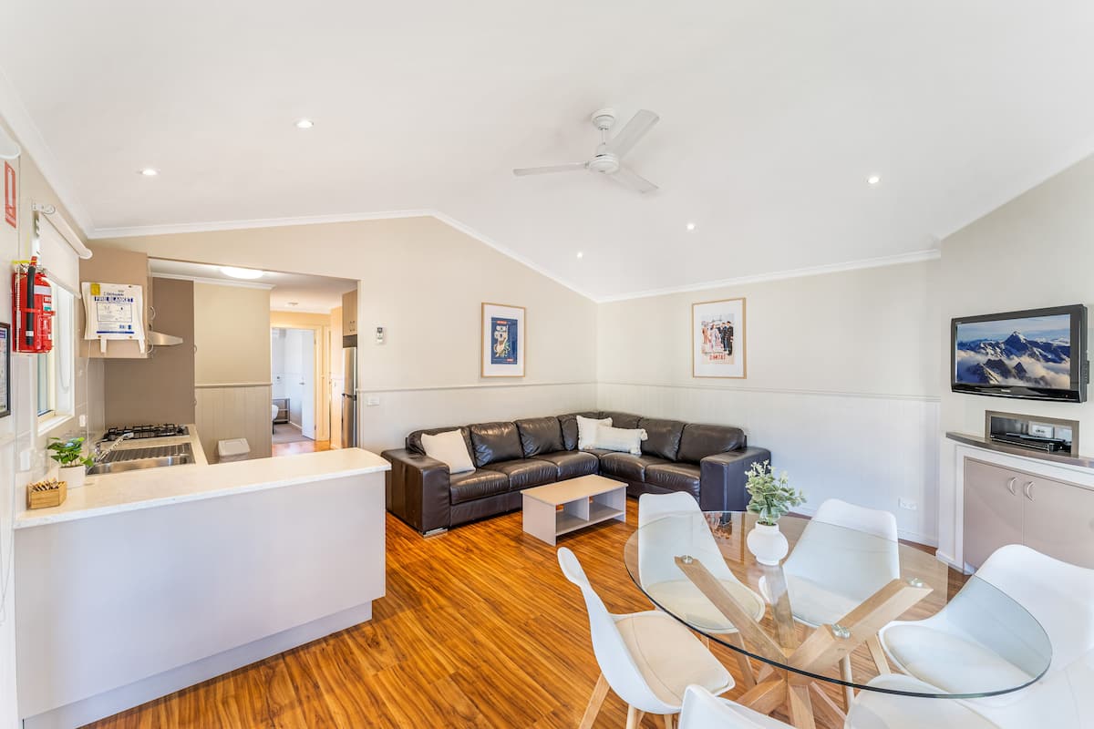 Ein modernes Wohnzimmer mit einer schwarzen Ledersofa, einem Glastisch und weißen Stühlen. Es gibt einen Küchenbereich mit einer weißen Arbeitsplatte und einem eingebauten Backofen. Ein Deckenventilator ist sichtbar, und der Boden ist mit Holz versehen.