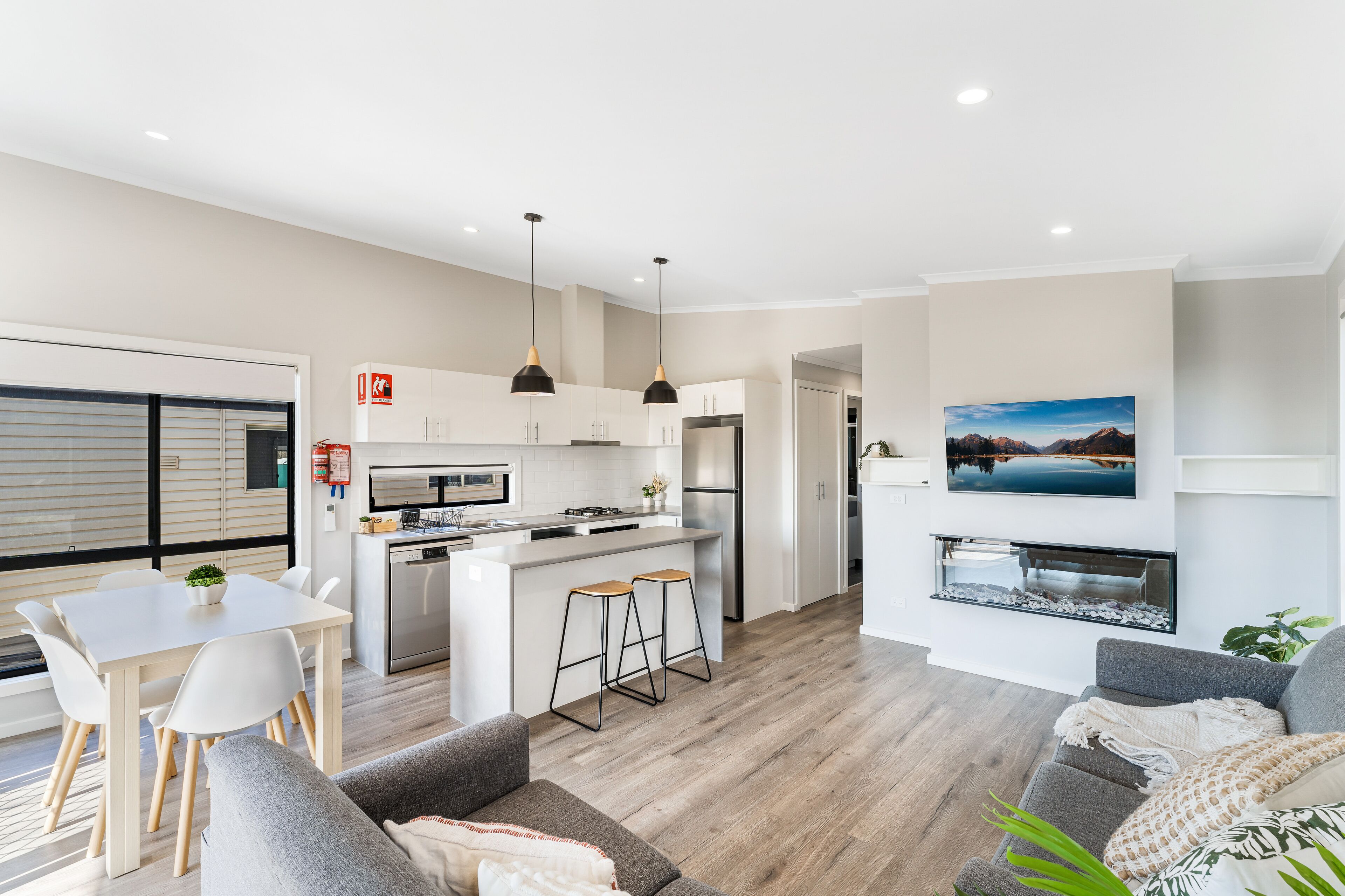 A modern kitchen with white cabinets, a central island, and bar stools. There is a dining area with a white table and chairs, and a gray sofa in the living room.