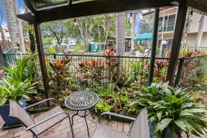 A covered outdoor seating area with chairs and a table, surrounded by various plants and a view of a building in the background. - Koala Court Holiday Apartments (Cairns North)