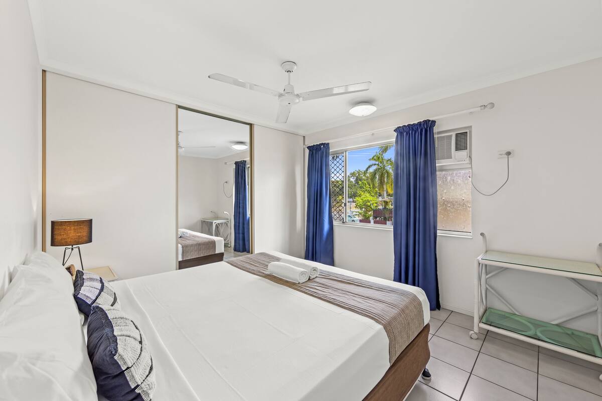 Une chambre d’hôtel équipée d’un lit, d’un bureau, d’un miroir, d’un ventilateur de plafond et d’une fenêtre avec des rideaux.