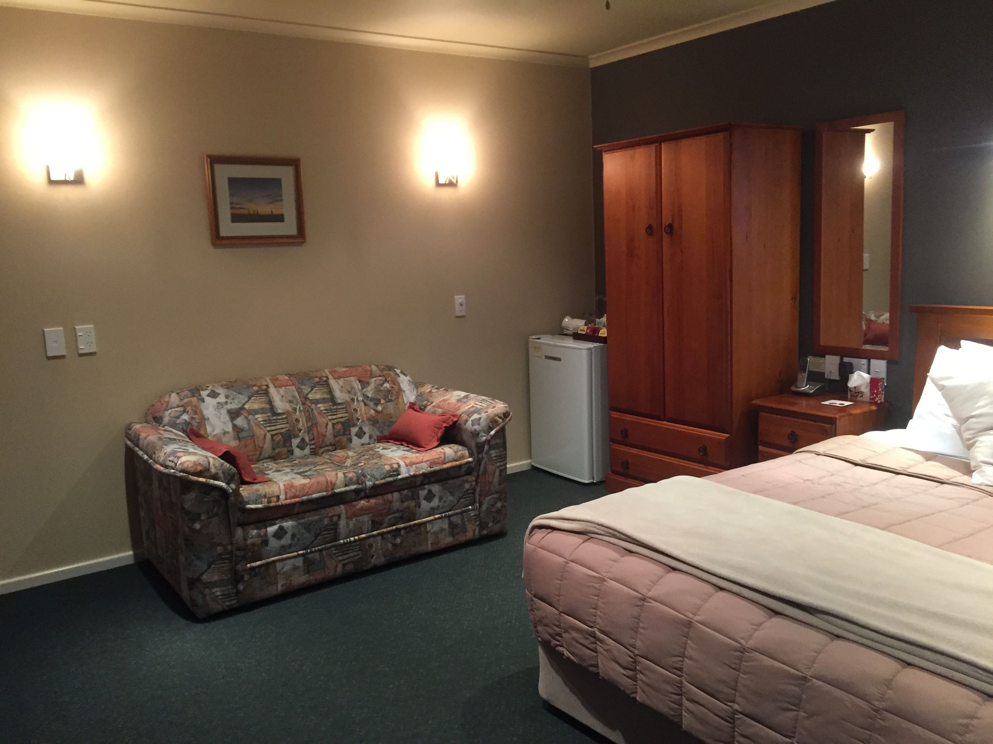 Une chambre à coucher comprenant un lit, un canapé à motifs, une armoire, un petit réfrigérateur et des lampes fixées au mur.
