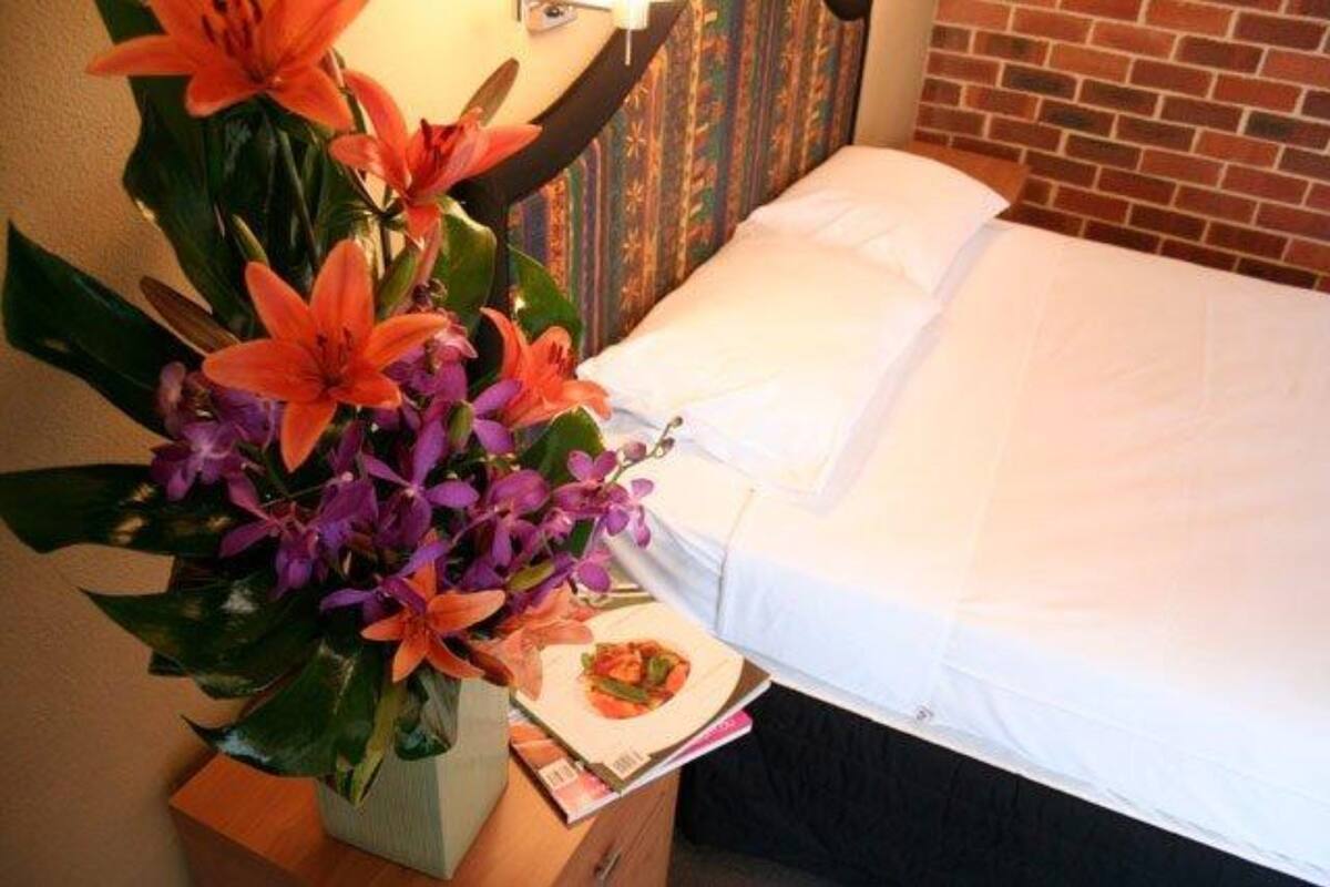 Una habitación de hotel con cama, una mesita de noche con un florero y un plato de comida.