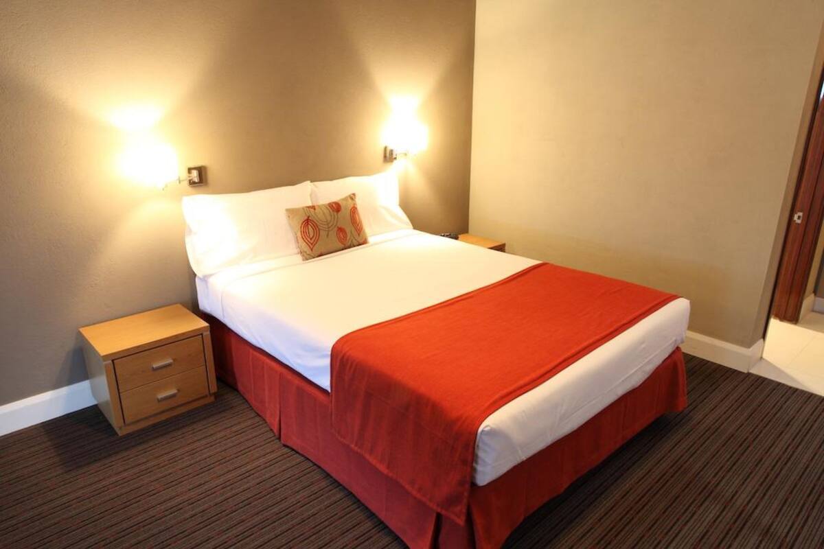 Una habitación de hotel con cama, mesita de noche y dos luces fijadas a la pared.