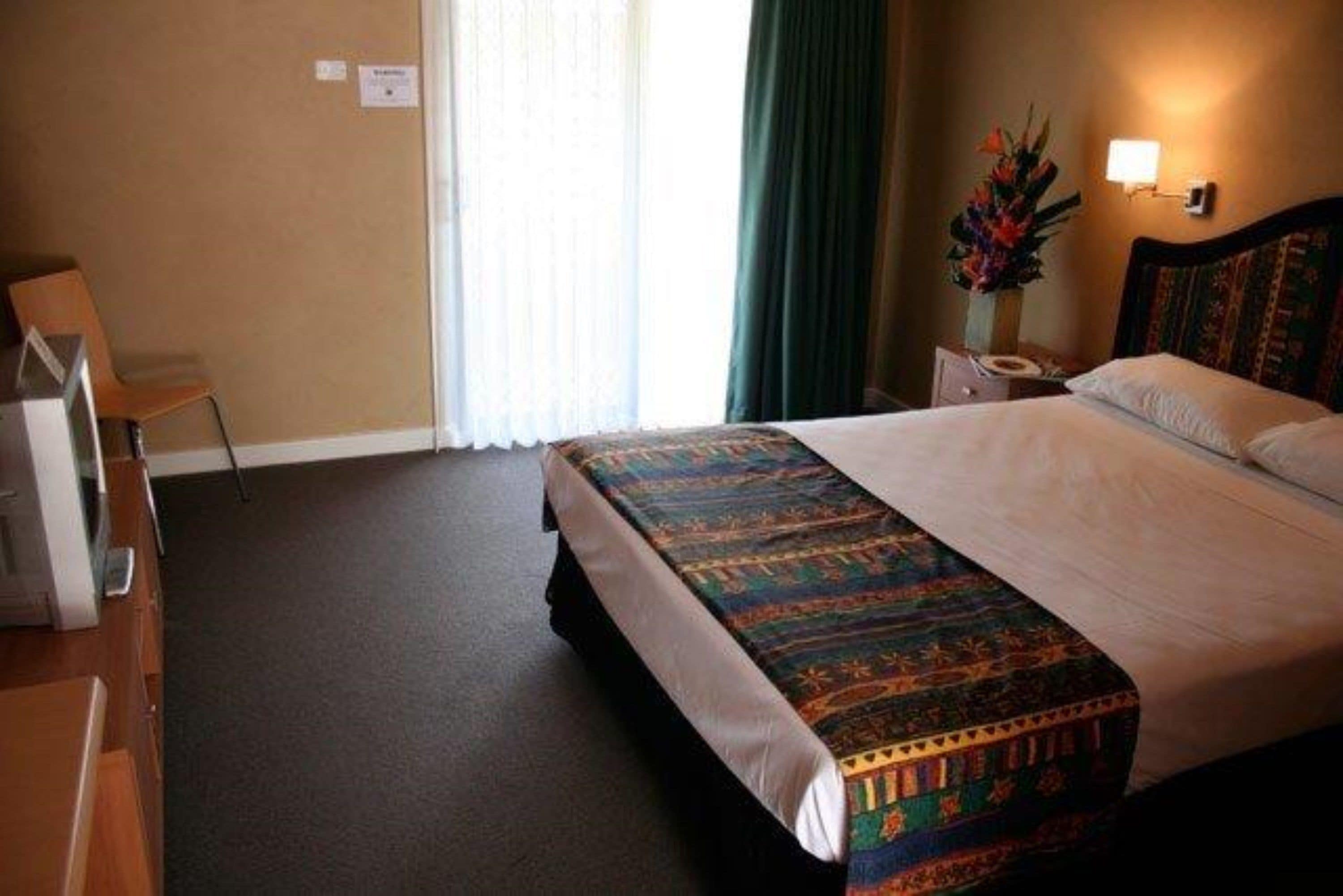 A hotel room with a large bed, a desk with a chair, a nightstand with a lamp, and a window with curtains.
