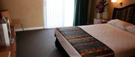 Habitación de hotel con una cama grande, un escritorio con silla, una mesita de noche con lámpara y una ventana con cortinas.