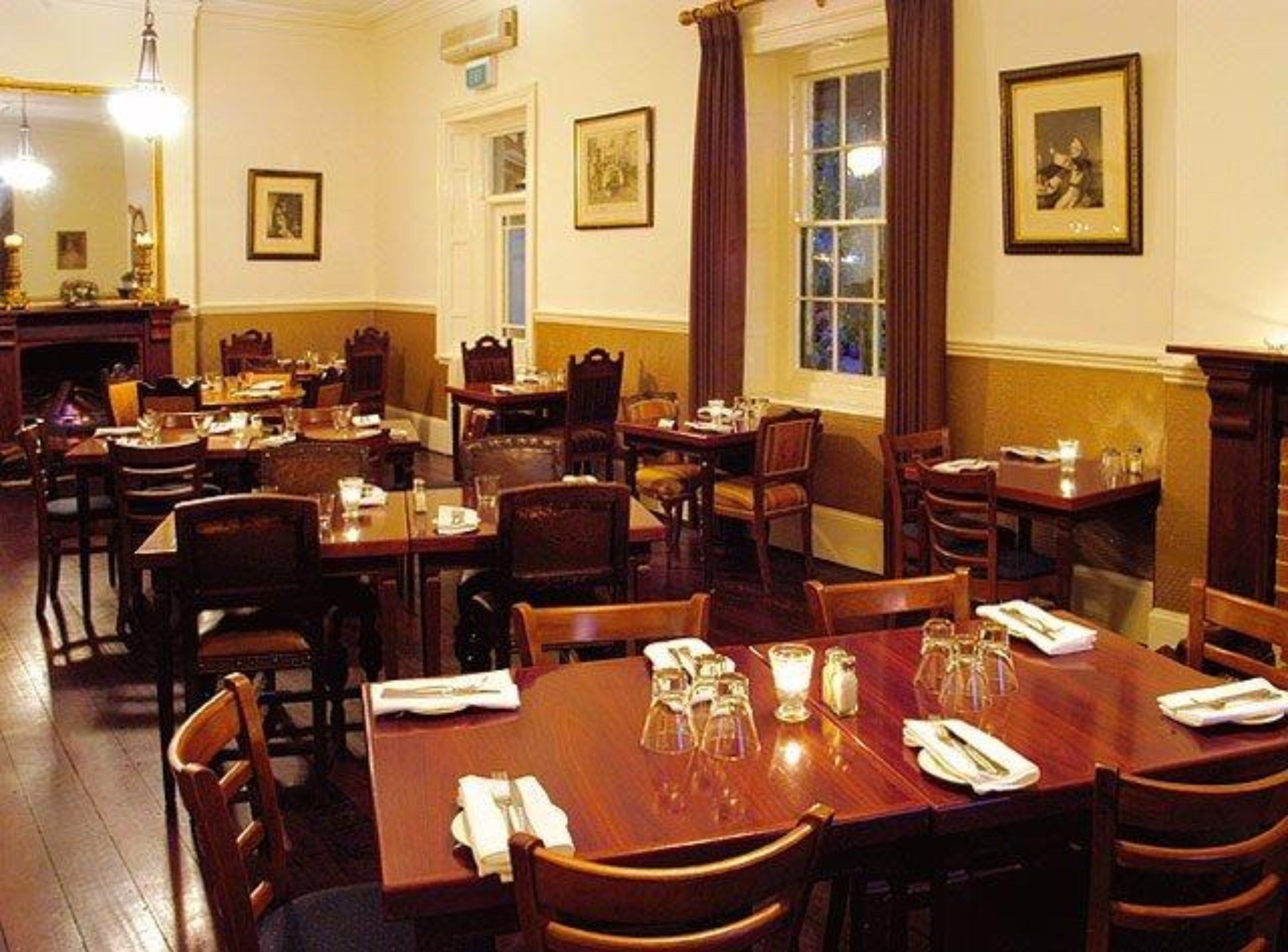 A dining area with multiple wooden tables and chairs, framed pictures on the walls, and a window with curtains.