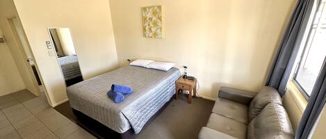 Habitación de hotel con cama, mesita de noche, silla, ventana con cortinas y una obra de arte colgada en la pared.