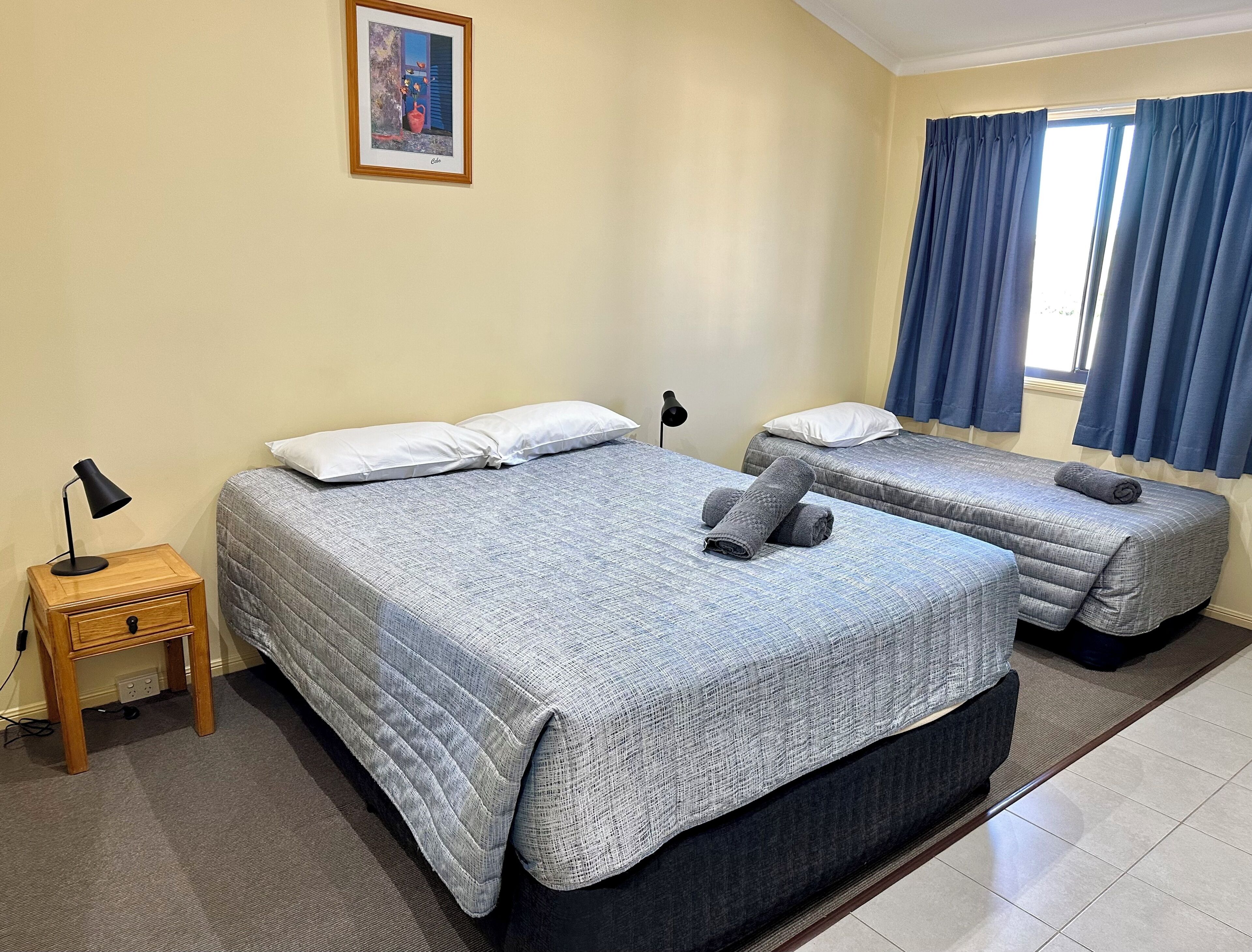 Une chambre d’hôtel avec deux lits, une petite table de chevet avec une lampe, un tableau au mur et une fenêtre avec des rideaux.