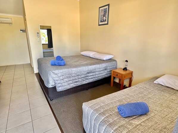 Quarto de hotel com duas camas, uma pequena mesa de cabeceira de madeira, um quadro na parede e uma janela com cortinas.