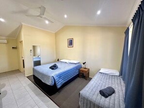 Quarto de hotel com uma cama grande, uma cama menor, uma mesinha de cabeceira, um ventilador de teto e uma janela com cortinas.