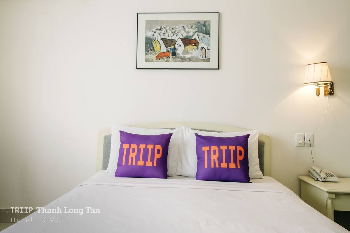 A hotel room with a bed, two purple pillows with the word "TRIP" on them, a nightstand with a lamp, and a framed picture on the wall.