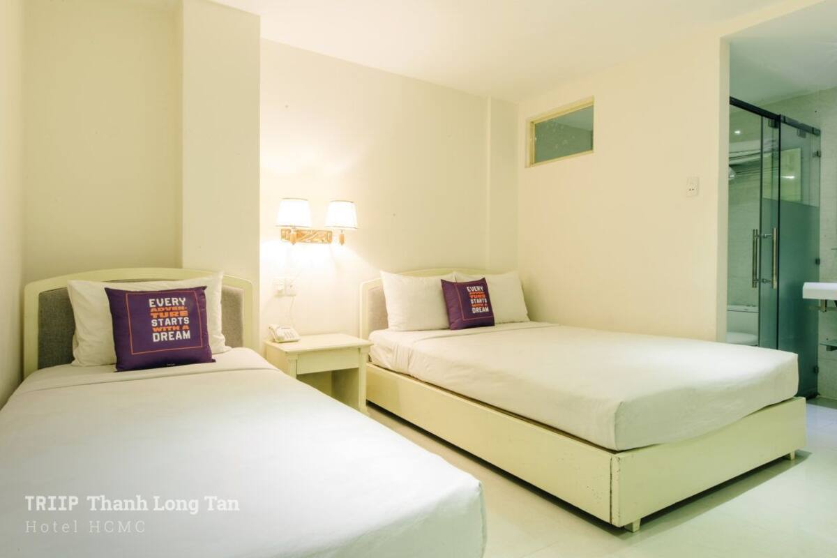 A hotel room with two beds, a nightstand with lamps, and a bathroom visible through a glass door.