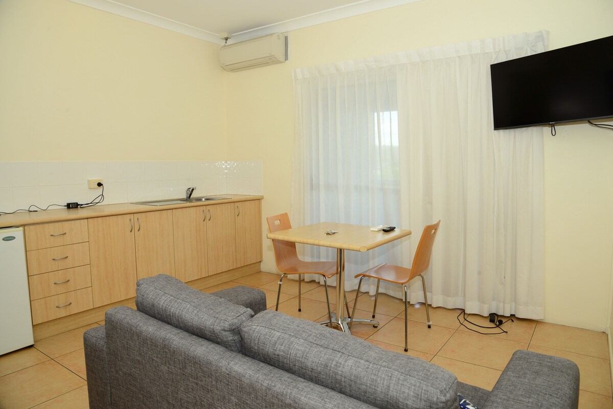 A compact living space with a kitchenette, a sofa set, a dining table with chairs, and a TV mounted on the wall.