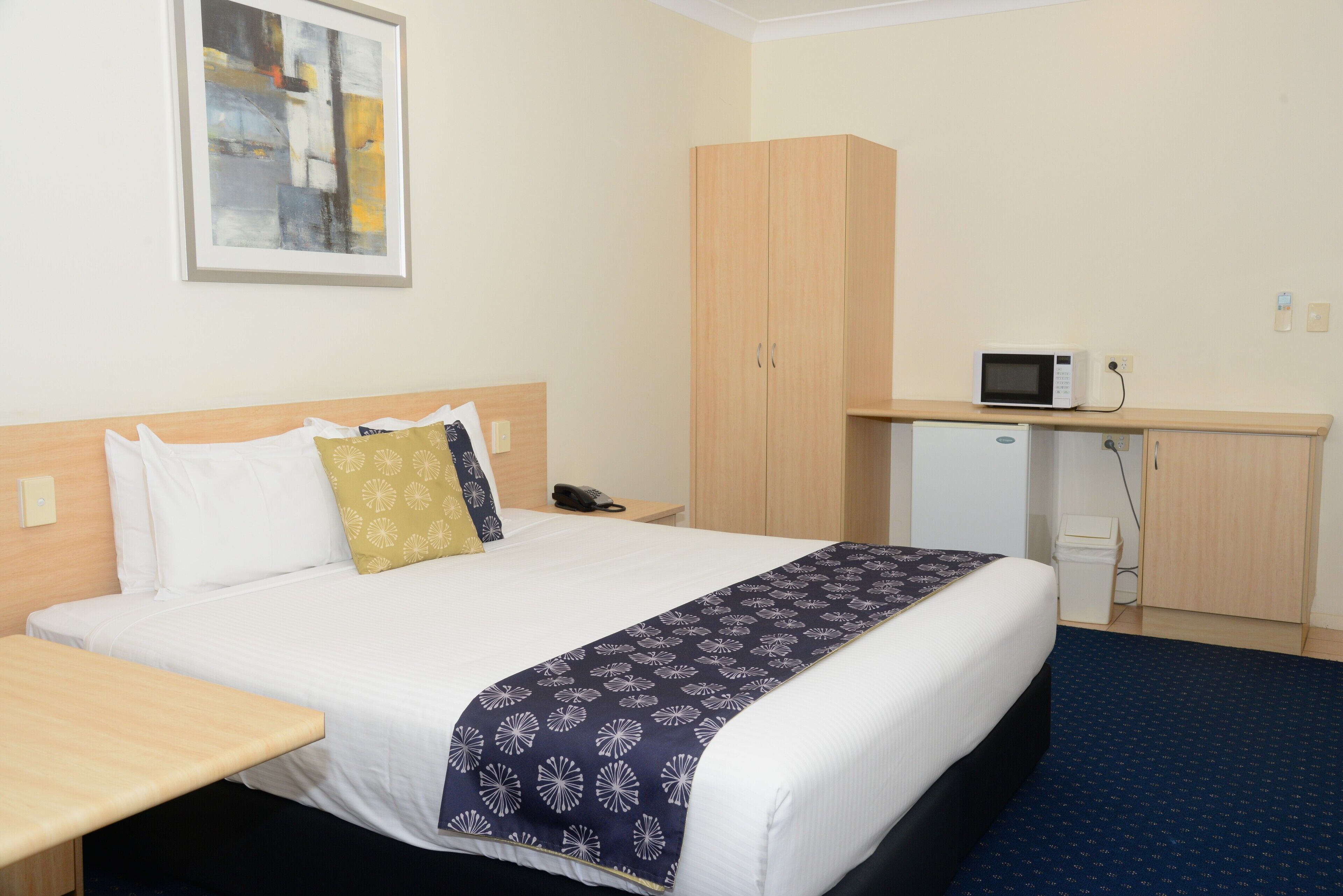 A hotel room with a bed, bedside table, a desk with a microwave, a telephone, and a cabinet.