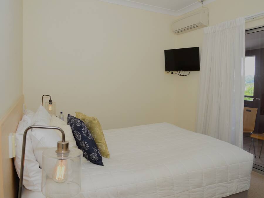 A hotel room with a bed, a bedside table with a lamp, a flat-screen TV mounted on the wall, and a window with curtains.