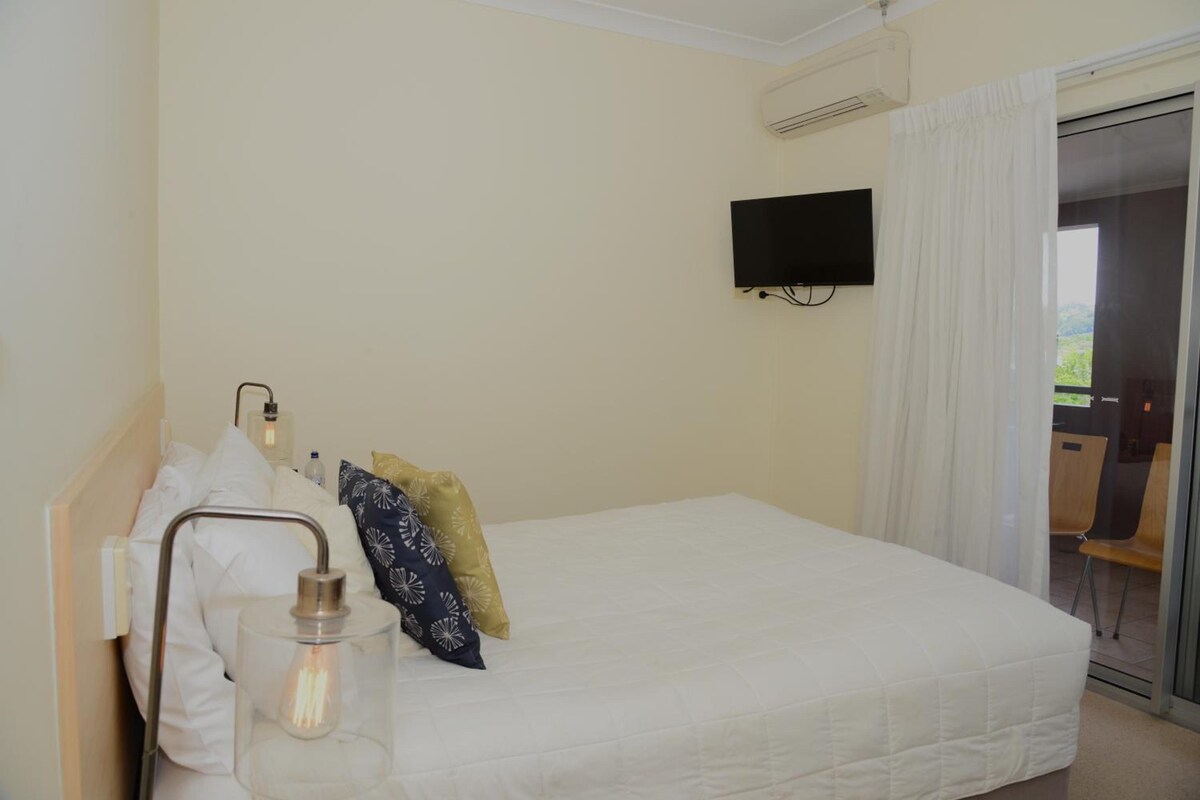 A hotel room with a bed, a bedside lamp, a television mounted on the wall, and a view of the outdoors through a door.