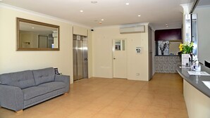 Et hotellobby med en grå sofa, en receptionsdisk, et spejl, en elevator og et skilt, der angiver mulighed for madkasse service.