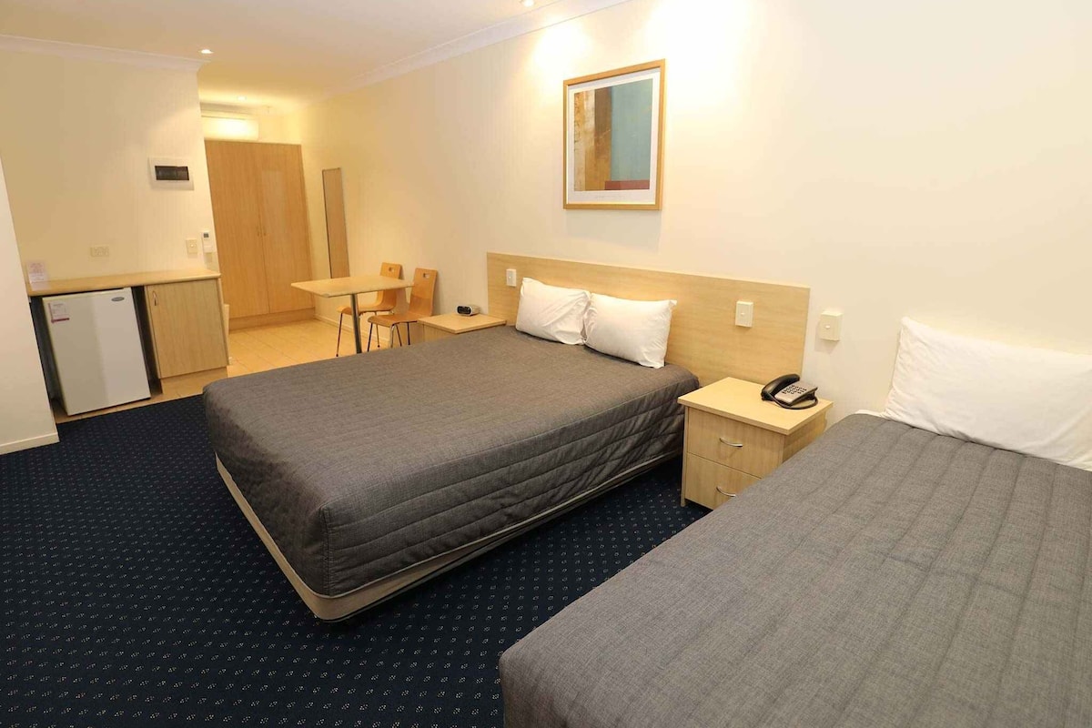 Quarto de hotel com duas camas, uma escrivaninha, uma cadeira, uma kitchenette pequena e um espelho na parede.