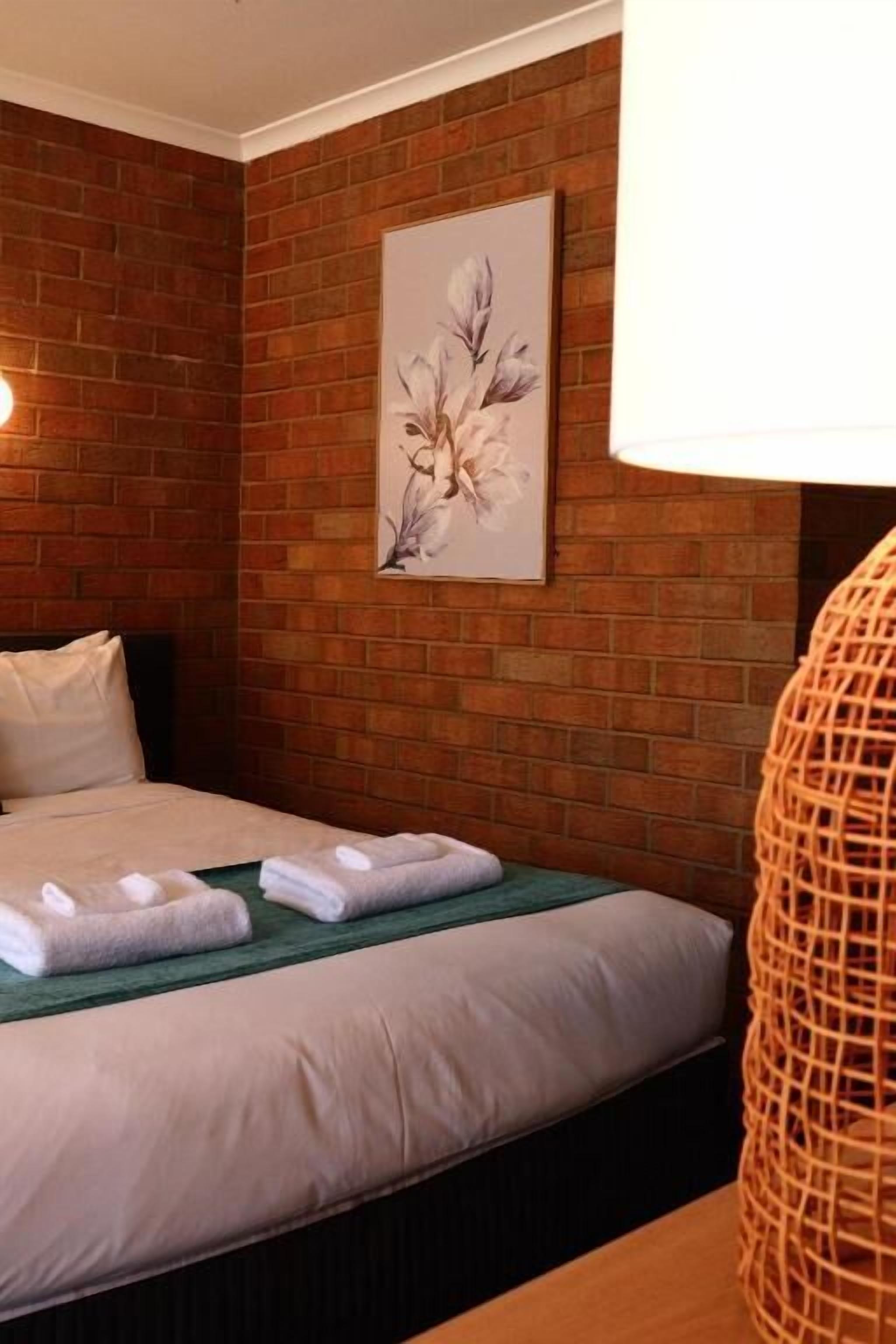 A hotel room with a brick wall, a bed with white linens, a green throw, and a framed floral artwork.