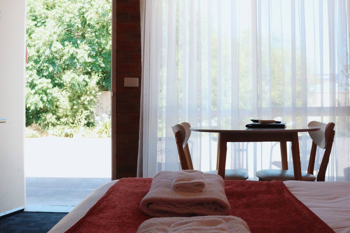 Une chambre d’hôtel avec un lit, une table et des chaises. Il y a une fenêtre avec des rideaux, et la vue donne sur un espace verdoyant.