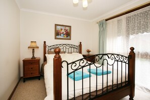 A bedroom with a wooden bed frame, a nightstand with a lamp, a framed floral painting on the wall, and a window with sheer curtains.